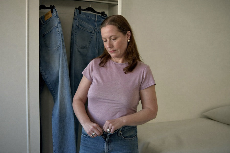 A woman at her closet in the morning, struggling with the waistband button of her jeans